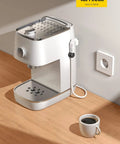 Compact white coffee machine with a cup of coffee on a wooden counter next to a wall plug.