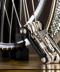All In One Bike Multitool Kit With Chain Breaker And Tire Repair in compact stainless steel design beside bicycle wheel.
