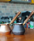 Two DCS Series Thickest Hammered Copper Turkish Greek Arabic Armenian Coffee Pot With Wooden Handle For Stovetop Brewing 20 Fl Oz Cezve Jezve Ibrik Briki Pro Grade on a table.