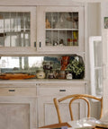 White kitchen cabinet with glass doors, displaying various decorative items and jars, open window alongside, wooden chair in foreground.