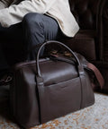 Carry On Ready Duffle Travel Bag With Clamshell Opening, 33 Liter Capacity, Pebble Full Grain Leather In Chocolate by a sofa