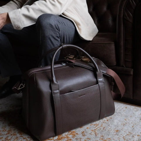 Carry On Ready Duffle Travel Bag With Clamshell Opening, 33 Liter Capacity, Pebble Full Grain Leather In Chocolate by a sofa