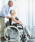 Elderly couple, man pushing woman in wheelchair, in a bright room with hardwood floors.