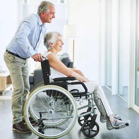 Elderly couple, man pushing woman in wheelchair, in a bright room with hardwood floors.