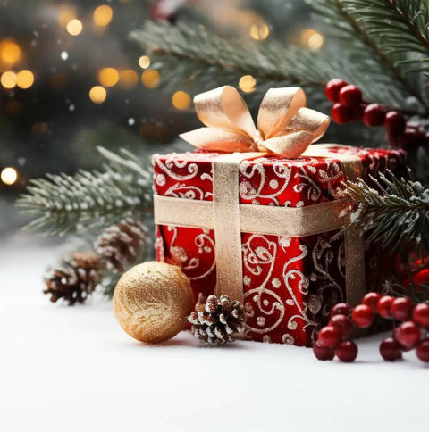 Festive Christmas gift box with gold ribbon, red ornament, and pine branches with snow, surrounded by holiday decorations.