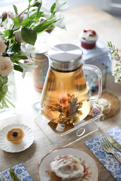 One-Touch 2-In-1 Tea Maker Teapot and Kettle with stainless steel filter lid, filled with tea, on a table setting with flowers.