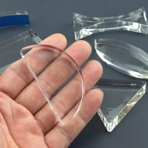 Hand holding semicircular lens from Six Piece Acrylic Glass Light Prism Set For Physics Lab Teaching, surrounded by other prisms and lenses.