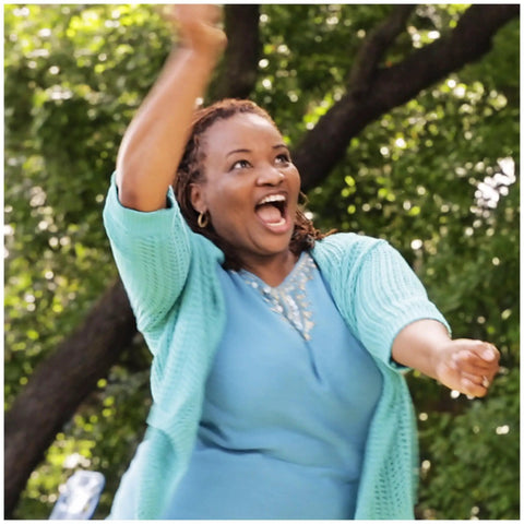 Person joyfully raising arms outdoors with green trees in background, wearing blue outfit, smiling exuberantly.