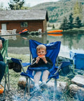 Child sitting in a blue folding camping chair by a lake, surrounded by nature, with houses in the background.