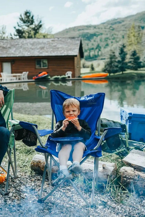 Child sitting in a blue folding camping chair by a lake, surrounded by nature, with houses in the background.