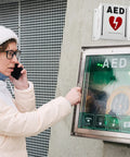 Front view of wall-mounted AED cabinet with glass door and green indicator; defibrillator visible inside.