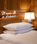 Glowing 'It was all a dream' neon sign above a bed with pillows and lamp, warm light ambiance in wooden room decor.