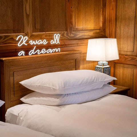 Glowing 'It was all a dream' neon sign above a bed with pillows and lamp, warm light ambiance in wooden room decor.