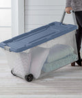 Person wheeling a clear heavy duty plastic container with snap lid, holding pillows and blankets, for home organization.