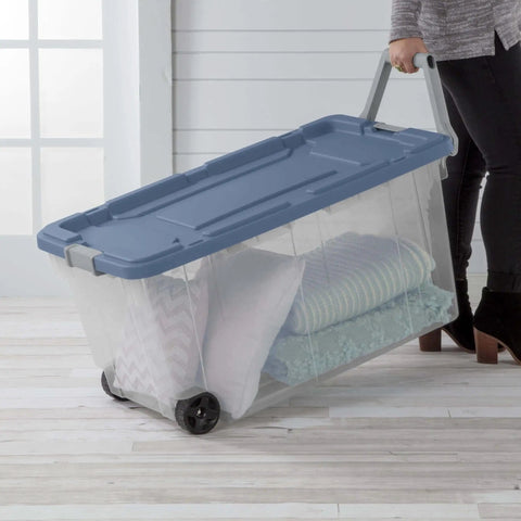 Person wheeling a clear heavy duty plastic container with snap lid, holding pillows and blankets, for home organization.
