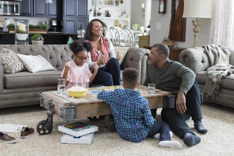 Family playing board game on carpeted living room floor, near beige sofa, emphasizing cozy home atmosphere.