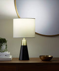Modern table lamp on wooden shelf with books, bowl, and mirror reflecting soft light