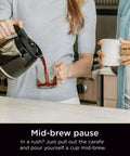 Person pouring coffee from a 12-cup programmable coffee maker with glass carafe into a cup, showcasing mid-brew feature.
