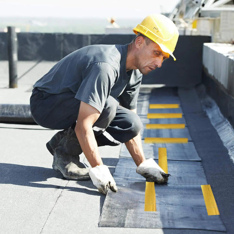 Construction worker on roof with roll of adhesive aluminum tape, wearing yellow hard hat, applying reflective barrier material