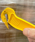 Disposable Film Cutter With Sharp Steel Blade And Safe Hidden Blade Design For Easy Cutting Of Shrink Wrap Stretch Wrap Tape And Plastic Straps Yellow Heavy Duty Office Tool For Warehouse And Mailroom being held against plastic wrap.