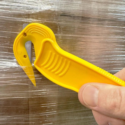 Disposable Film Cutter With Sharp Steel Blade And Safe Hidden Blade Design For Easy Cutting Of Shrink Wrap Stretch Wrap Tape And Plastic Straps Yellow Heavy Duty Office Tool For Warehouse And Mailroom being held against plastic wrap.