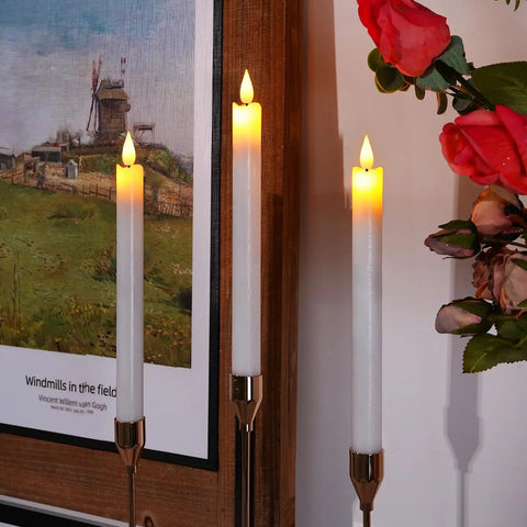 Three Eight Inch Ivory Flameless Taper Candles With Remote And Timer, with realistic flickering flame next to a framed painting.