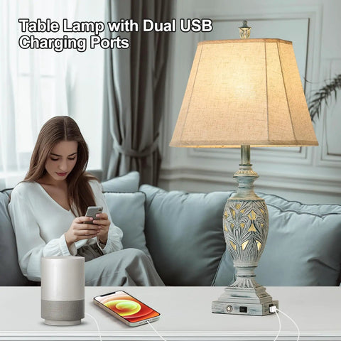 Woman using phone beside table lamp with dual USB charging ports, linen shade and detailed base; cozy living room setting.