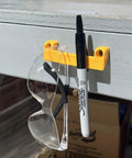 Yellow Rugged Tape Measure Holder For Garage Organization, Keeps Tools Handy On Ladder Or Workbench, Grab-And-Go Quick Install Tool Hook Pack Of 4, holding a marker and glasses on workbench.