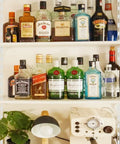 Bar shelf with assorted liquor bottles and decor items, including a plant and a coffee maker, on white wooden shelves.