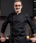 Heavy Duty Chef Coat With Chest And Sleeve Pocket Plus Hella Breathable Fabric That Keeps You Cool In The Heat worn by a middle-aged male chef in a modern kitchen.