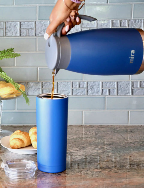 Pouring coffee from Stainless Steel Thermal Carafe Dispenser Vacuum Insulated Dual Wall Bottle For Hot Coffee Tea Or Cold Water 1.5 Liter 50oz Keeps Drinks Hot 12 Hours Cold 24 Sweat Free Admiral Blue into a blue tumbler on a kitchen counter.