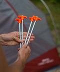 Hands holding Beefy 10-Inch Steel Tent Stakes Heavy-Duty Ground Anchors 4-Pack for camping with orange tops in front of a tent.