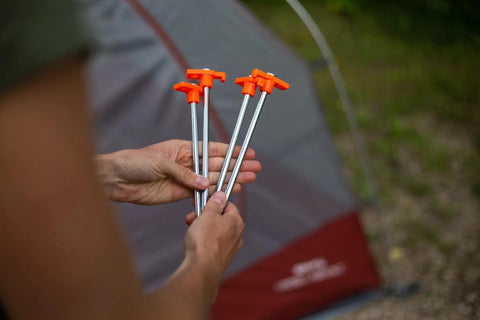 Hands holding Beefy 10-Inch Steel Tent Stakes Heavy-Duty Ground Anchors 4-Pack for camping with orange tops in front of a tent.