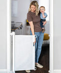 Woman carrying baby using Retractable Baby Gate For Stairs And Doorways, Extra Wide Mesh Barrier Extends Up To 71 Inches, 33 Inches Tall, Safe For Kids And Pets, Indoor Outdoor Use, One Handed Operation, Space Saving White Gate in doorway.