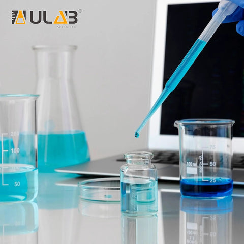 Close-up of ULAB Sterile Transfer Pipette dispensing blue liquid into a small glass bottle; lab glassware in background.
