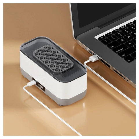 Portable Ultrasonic Cleaner With Rechargeable Battery For Jewelry And Watches on a desk, charging via USB next to a laptop.