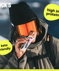 Person in winter gear eating Protein Bar Variety Pack With Candy Bar Taste, Low Calorie, Low Carb Keto Friendly Snack, snowy background.