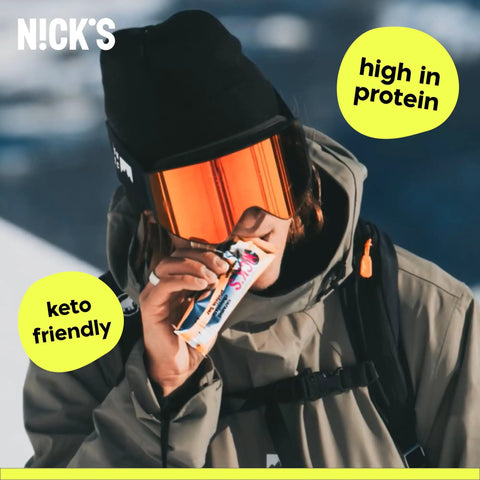 Person in winter gear eating Protein Bar Variety Pack With Candy Bar Taste, Low Calorie, Low Carb Keto Friendly Snack, snowy background.