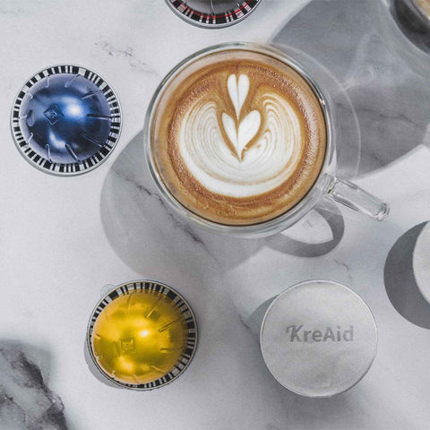 Top view of reusable aluminum foil lids for Nespresso VertuoLine pods next to brewed coffee and VertuoLine capsules on a marble surface.