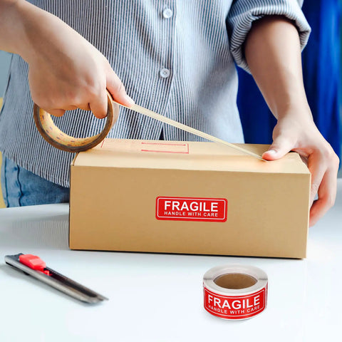 Person applying tape on box with Red Fragile Stickers, 150-Count Self-Adhesive Paper Labels For Packing And Shipping, Handle With Care Warning Labels For Boxes, Luggage, Glass, Cardboard, Bags, Moving Like A Pro.