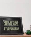 Digital Atomic Clock With Outdoor Temperature And Moon Phase Auto Time Sync Large Two Inch Digits Twelve Or Twenty Four Hour Display DST Auto Seven Time Zones No Fuss Setup Black Wall Or Desk Clock For Home And Office on wooden surface with plants.