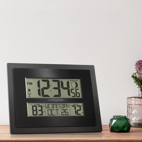 Digital Atomic Clock With Outdoor Temperature And Moon Phase Auto Time Sync Large Two Inch Digits Twelve Or Twenty Four Hour Display DST Auto Seven Time Zones No Fuss Setup Black Wall Or Desk Clock For Home And Office on wooden surface with plants.