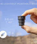Hand holding a small black two-pack portable bidet nozzle for travel, fitting any bottle, eco-friendly personal hygiene accessory.
