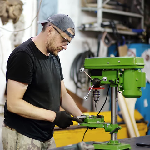 Person operating a green industrial drill press in a workshop setting with various tools and machinery in the background.