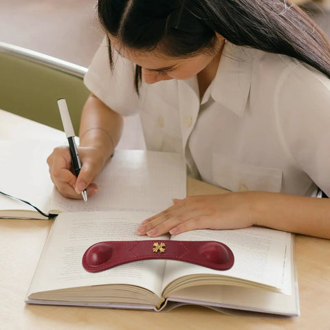 Red Leather Weighted Bookmark With Bronze Accent Corner Page Holder on an open book, used by a person writing notes.