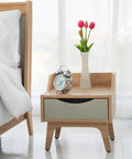 Wooden nightstand with a silver clock and pink flowers in a vase, beside a bed with white linens.