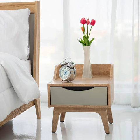 Wooden nightstand with a silver clock and pink flowers in a vase, beside a bed with white linens.