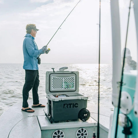 Person fishing on a boat with a 52 Quart Ultra Light Wheeled Cooler Insulated Ice Chest For Beach Camping Fishing Picnics And Boating With All Terrain Wheels Easy Push And Pull Mobility Premium Ice Retention 30 Percent Lighter Than Rotomolded Coolers Olive.