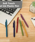 Assorted colors of soft touch gel ink pens placed on a wooden desk near a laptop and a plant, showcasing stationery essentials.