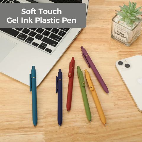 Assorted colors of soft touch gel ink pens placed on a wooden desk near a laptop and a plant, showcasing stationery essentials.